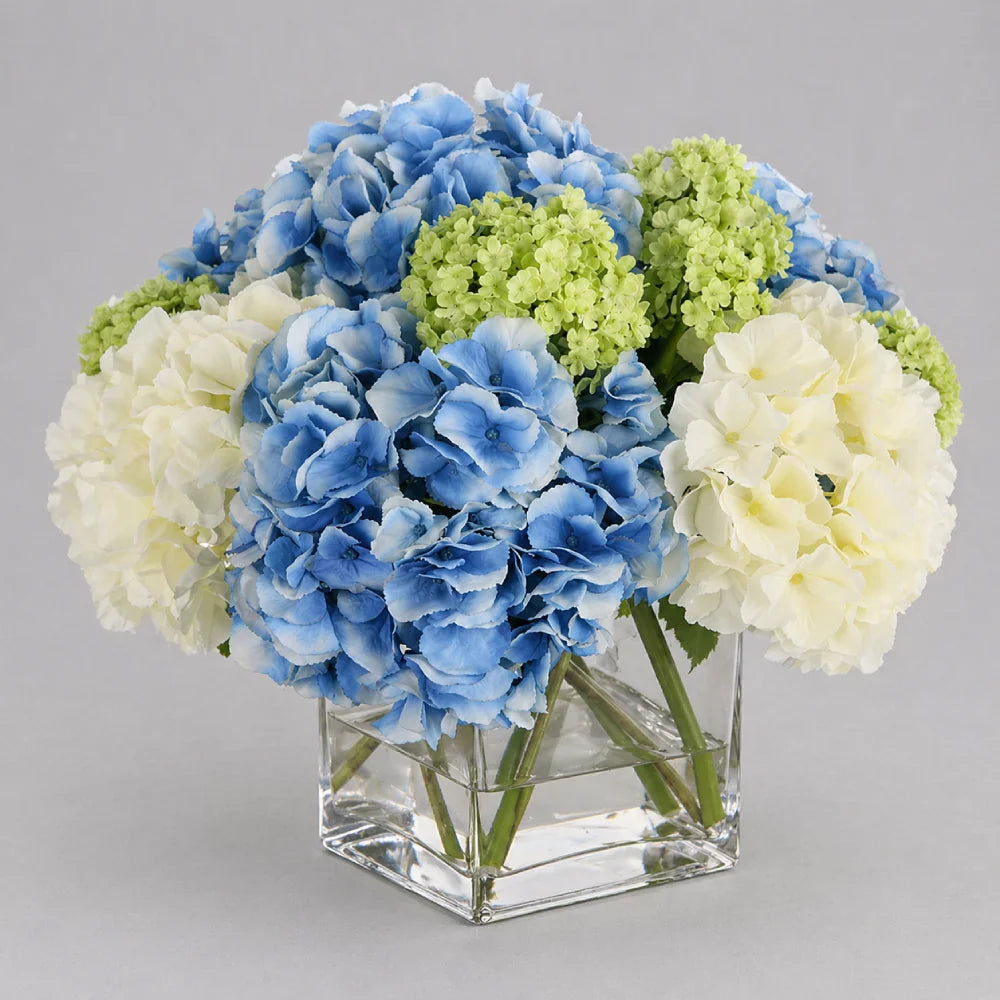 Square glass vase with blue, green, and white hydrangeas on a gray background