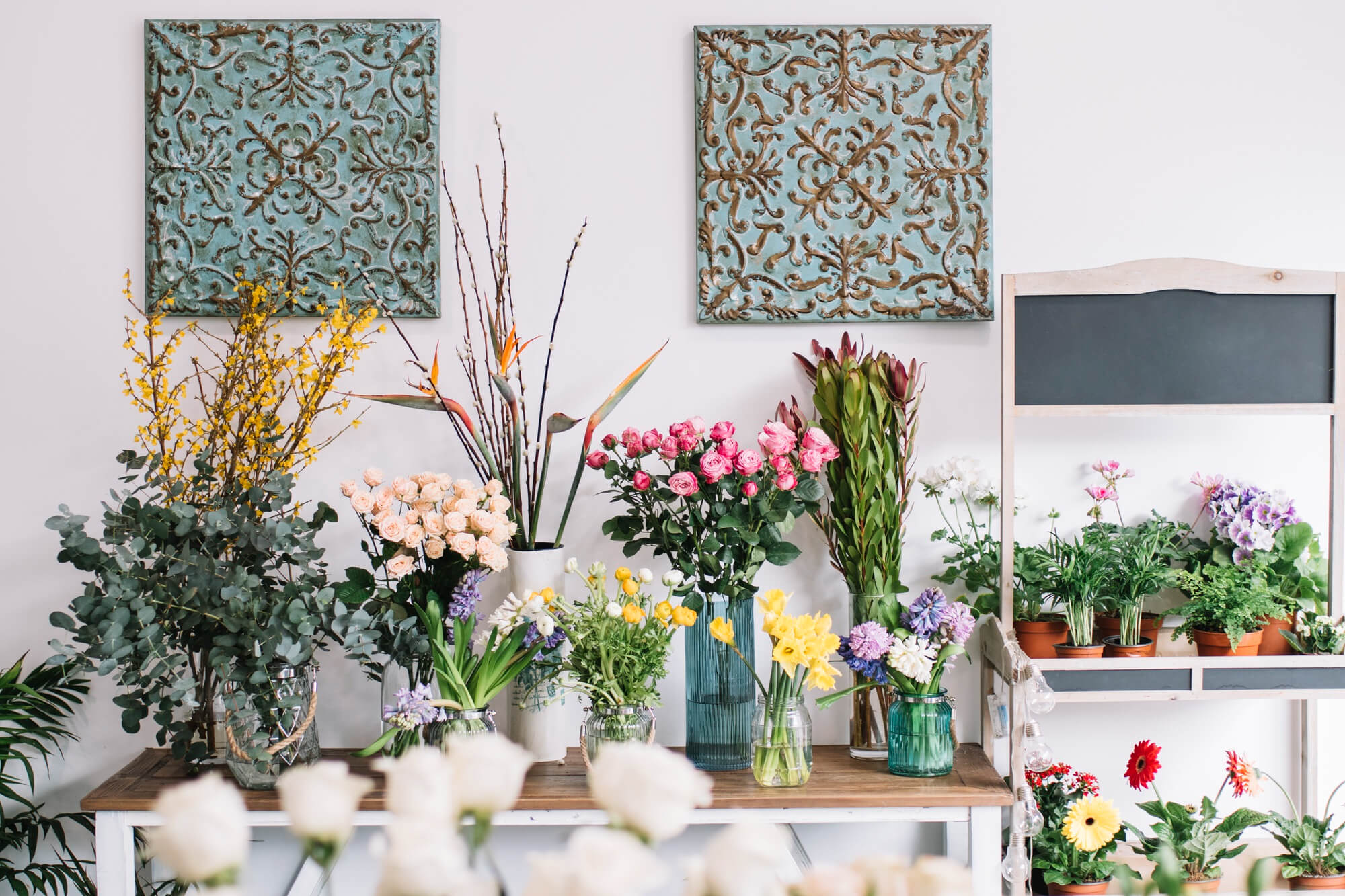 Floral arrangements in various vases on a wooden table with decorative wall art in the background.