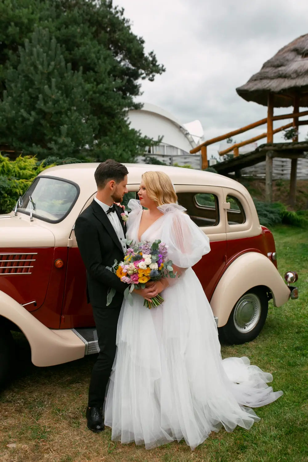 Couple in wedding attire standing in front of a vintage car with a scenic background.