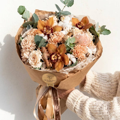 Bouquet of flowers wrapped in brown paper with a visible brand label, held by a person wearing a beige sweater.