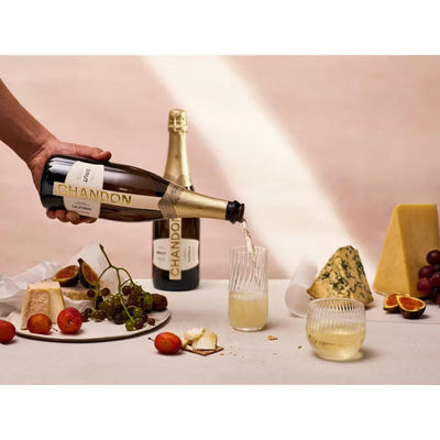 Chandon champagne being poured into a glass with a plate of cheese and fruit on a table.