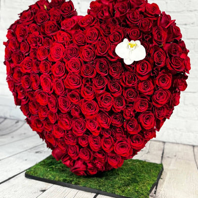 Heart-shaped arrangement of red roses with a white flower in the center on a wooden floor.