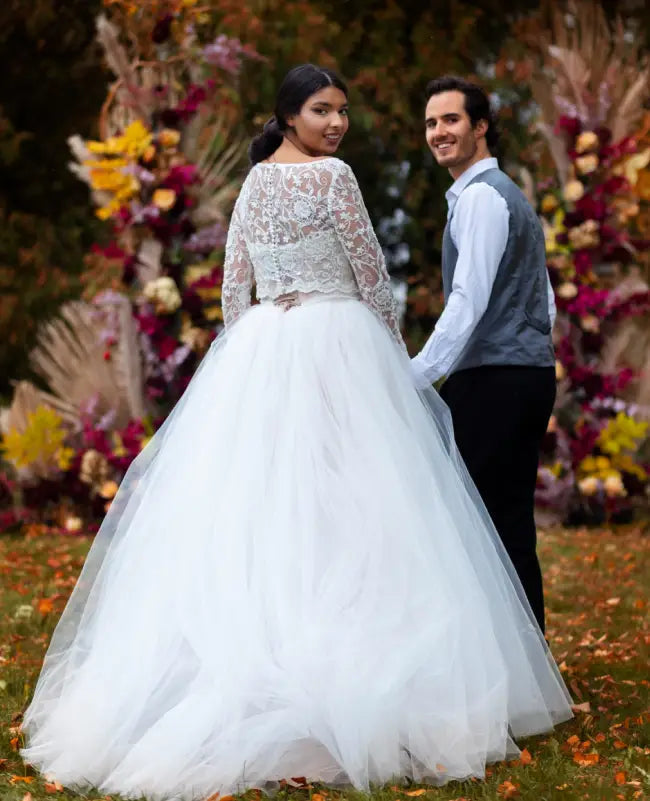 Woman in a white wedding dress standing next to a man in formal attire with a floral and autumn-themed background.