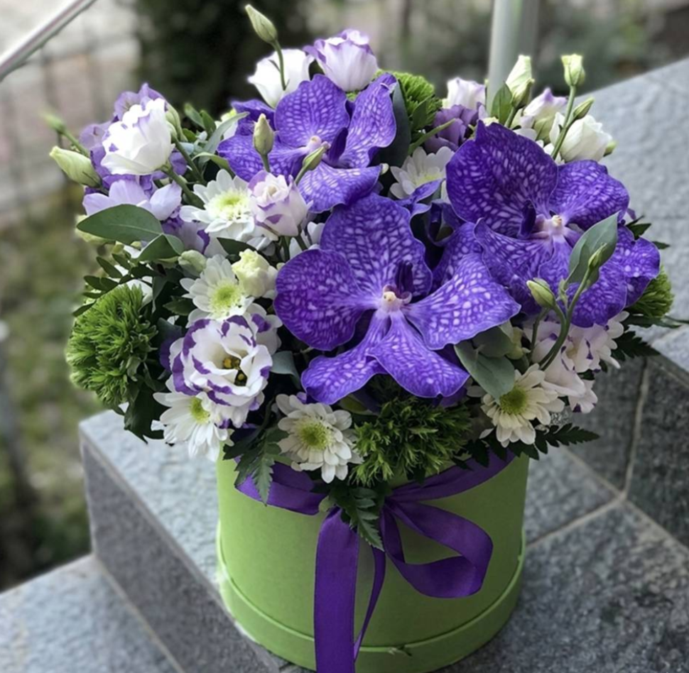 Bouquet of purple and white flowers in a green box with a purple ribbon on a stone surface.