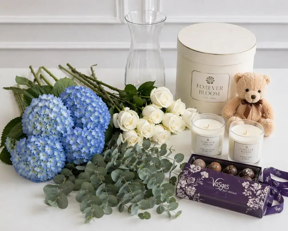 Floral arrangement with blue hydrangeas, white roses, and eucalyptus next to candles and a teddy bear on a white surface.