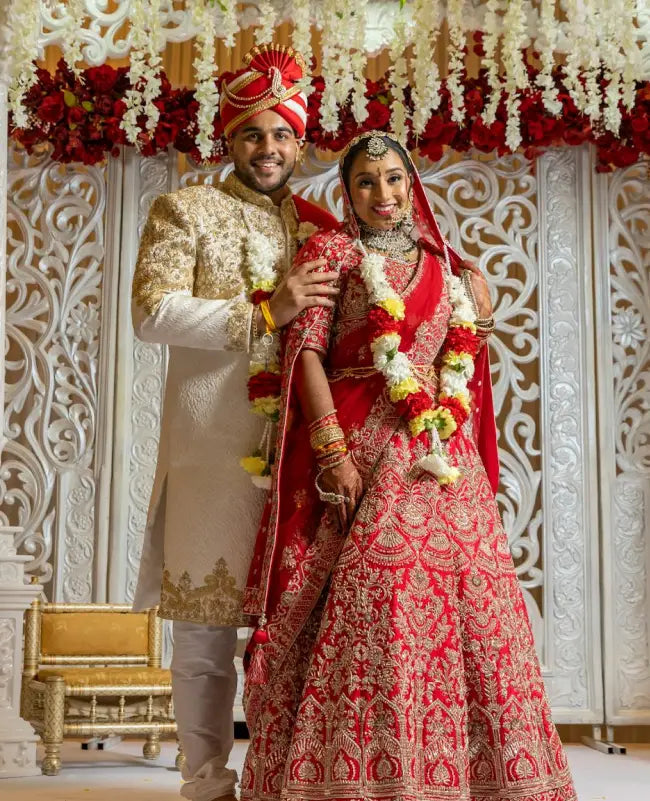 Couple in traditional wedding attire with floral decorations and intricate wall patterns.