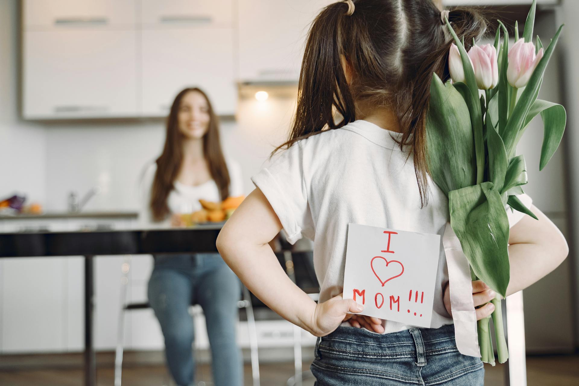 Person holding tulips and a card with 'I love mom' in a kitchen setting