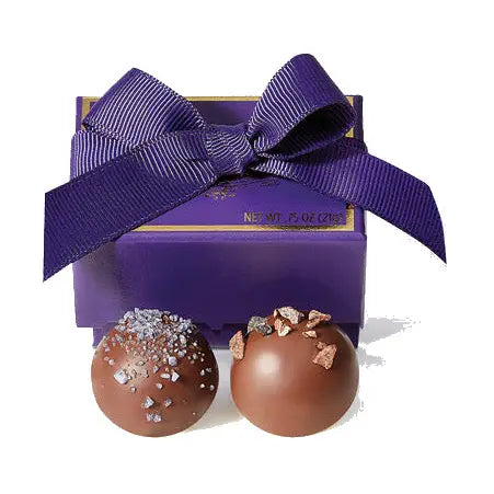 Two chocolate truffles with decorative toppings in front of a purple gift box with a bow.